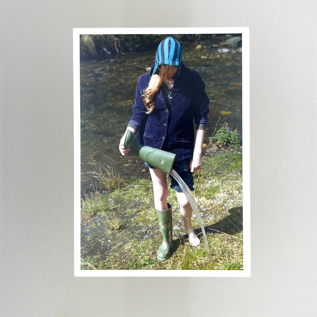 Postcard of a woman wearing a striped blue hoodie under a blue jacket, trousers rolled up, with one wellington boot. She's walking next to a river and pouring water out of the other wellington boot that's she's holding. 
