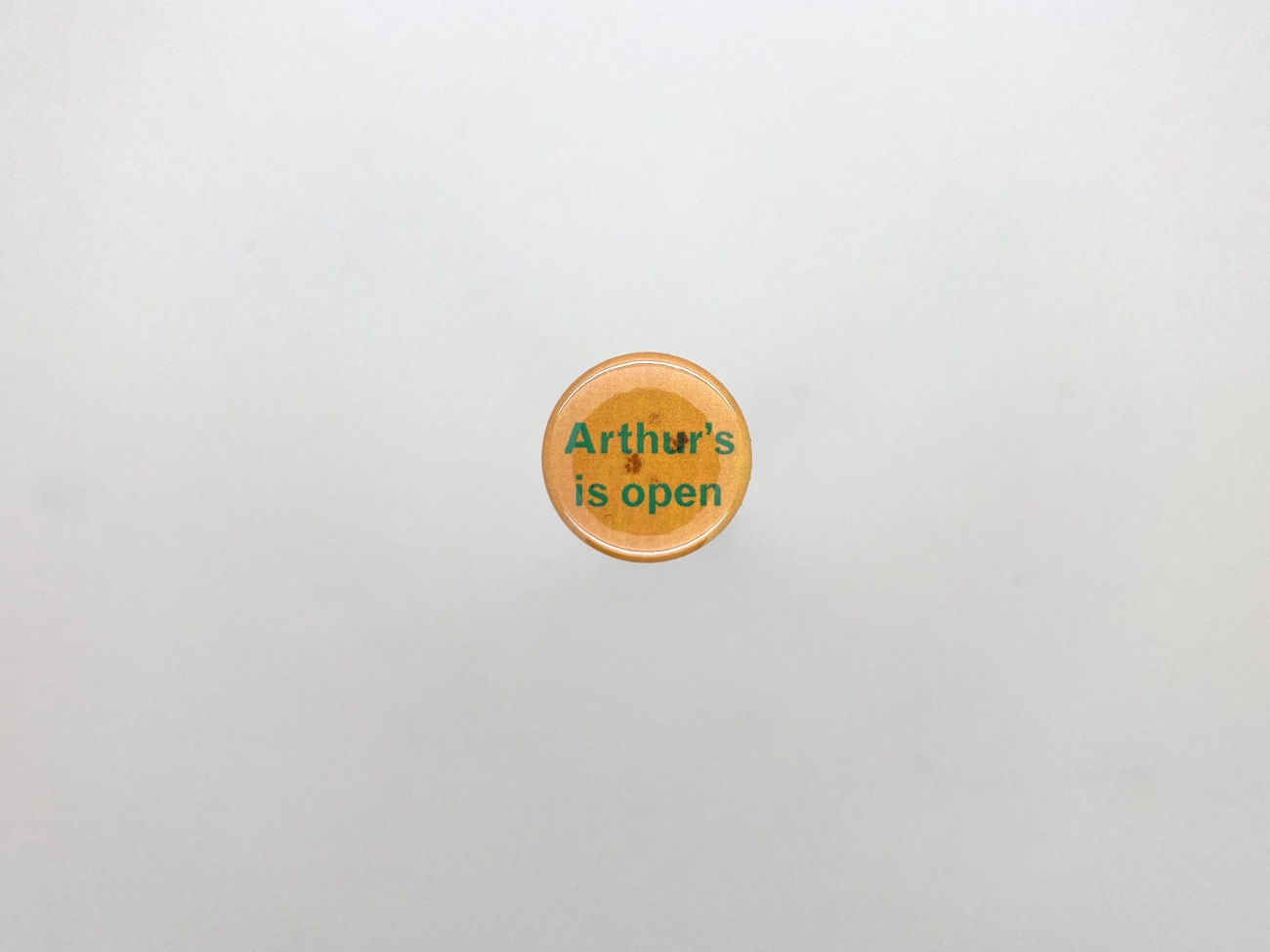 A photo of a small 10p piece orange badge with the words Arthur's is open 