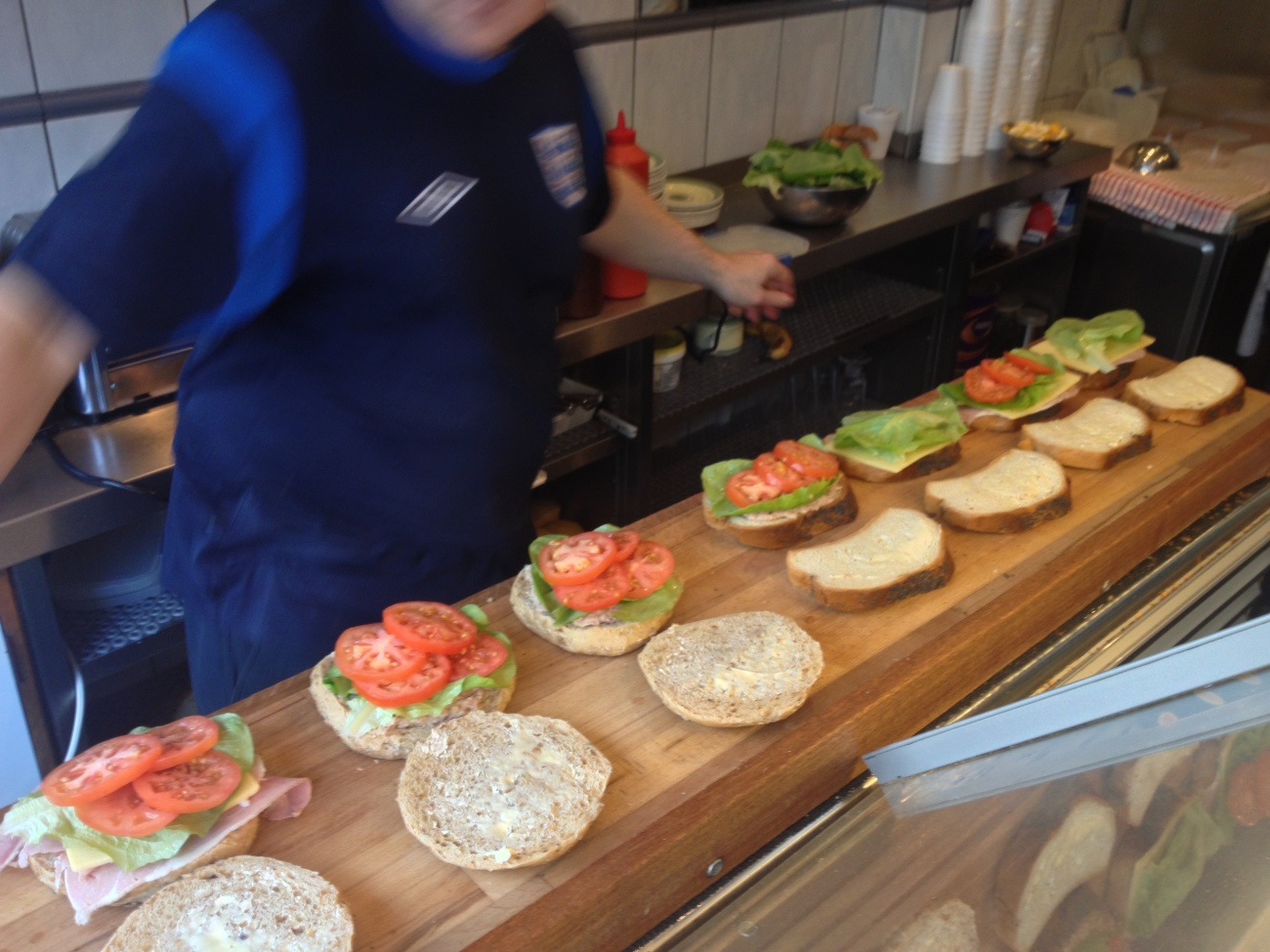 A blurred James speeds along as he adds individual ingredients to the 7 open baps and sandwiches. All but two have tomato. All have lettuce. 